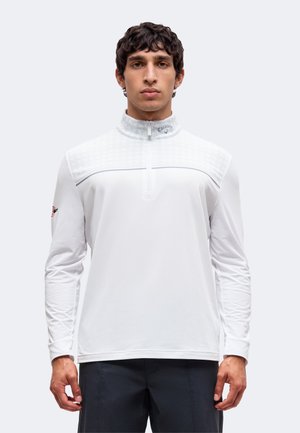 Jeune homme aux cheveux bouclés foncés portant une chemise de golf blanche à manches longues avec un col zippé et un pantalon foncé, regardant devant avec une expression neutre.