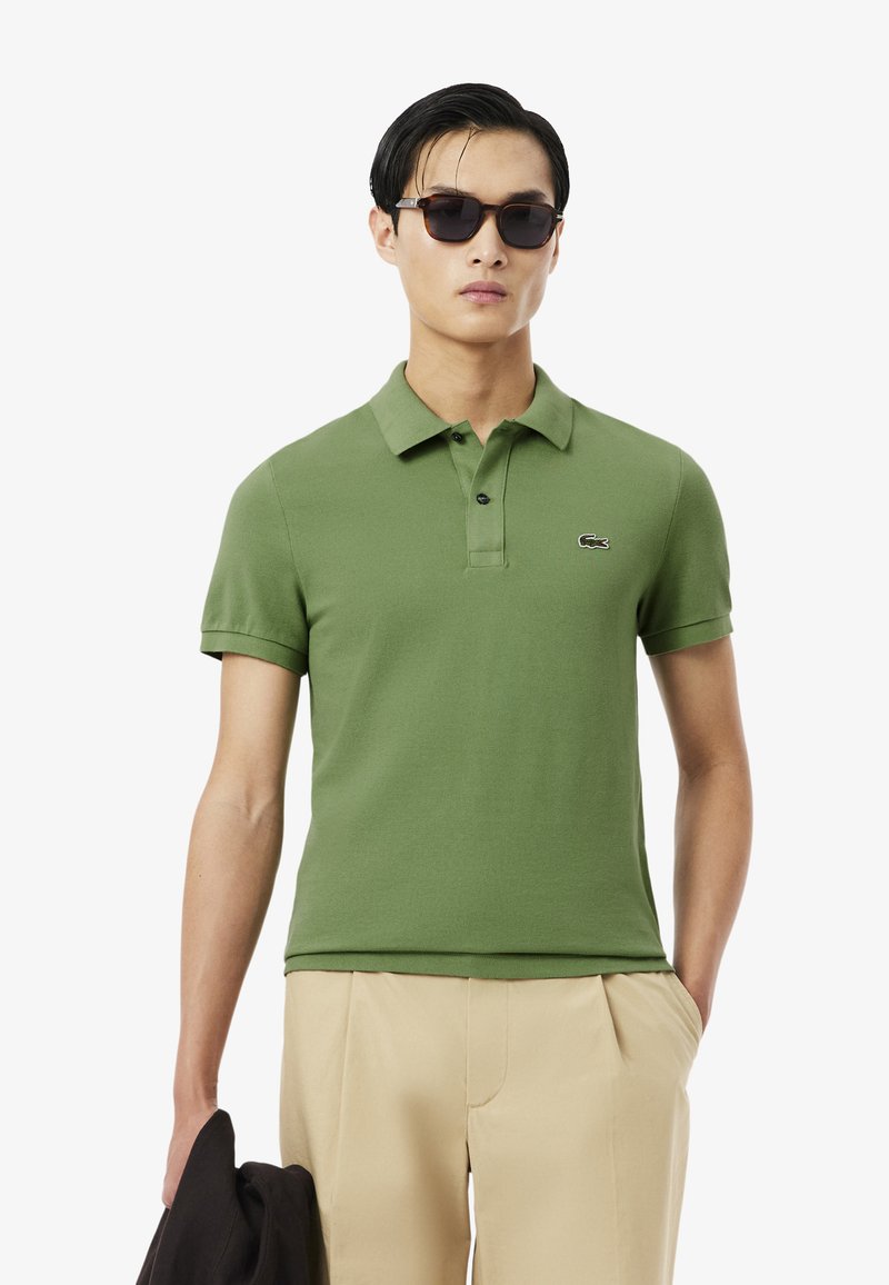 Mann trägt dunkle Sonnenbrille, grünes Poloshirt mit kleinem Logo, beige Hose, hält eine dunkle Jacke in der Hand, steht vor einfarbigem Hintergrund.