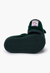 Zapato infantil verde aterciopelado con suela de goma texturizada, que presenta una etiqueta cosida con un diseño de mono y el texto "Hecho en España".