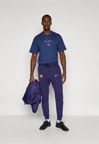 Herenoutfit met een marineblauw t-shirt met de tekst "BARÇA", paarse joggingbroek met logo en zwarte sportschoenen. Inclusief een paarse jas.