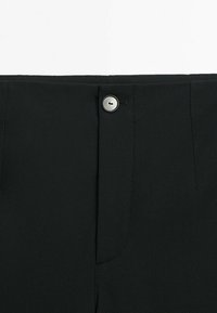 Gros plan sur la ceinture d'un pantalon noir avec un bouton argenté et des coutures visibles autour de la braguette et des poches.