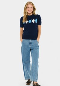 Leende kvinna med händerna i fickorna, iklädd en mörkblå argyle kortärmad tröja, ljusblå vida jeans och bruna loafers på vit bakgrund.