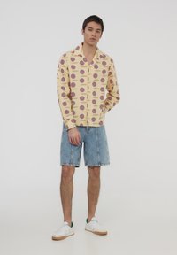 Maglietta gialla a maniche corte con motivo geometrico blu e rosso, abbinata a pantaloni corti di denim e sneakers bianche con dettagli verdi.