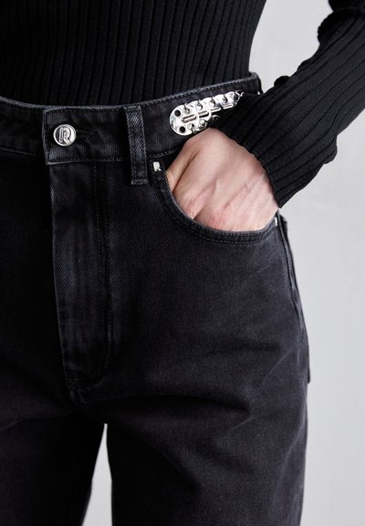 Black denim jeans with a sleek design, featuring a unique silver hardware accent at the waist and a front pocket with a visible hand.