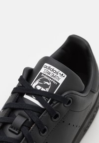 Sneaker Adidas Stan Smith noire avec une tige en cuir lisse, dotée de lacets noirs, d'accents perforés et d'une étiquette de marque sur la languette.