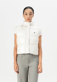 Beige puffer vest med høy krage og trykknapplukking, med quiltede seksjoner og logo-detalj, kombinert med grå bukser.