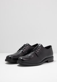 Chaussures habillées en cuir noir avec une texture lisse, bout arrondi, fermeture à lacets et un petit talon empilé. Présente des éléments de design minimalistes.