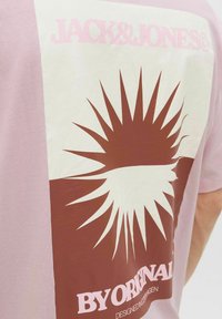 Lys pink T-shirt med et stort brunt grafik med et solstråledesign. Teksten inkluderer "JACK&JONES" og "BY ORIGINAL" i pink på en cremefarvet baggrund.