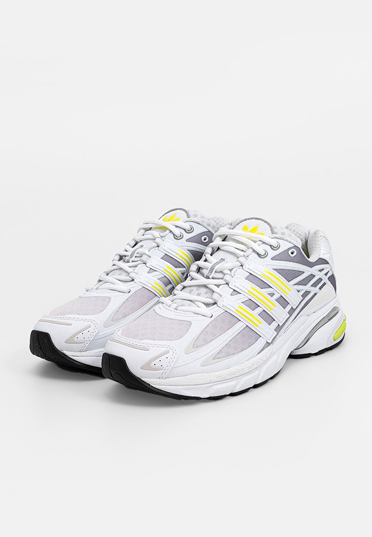 Zapatillas deportivas blancas con detalles en gris, parte superior de malla y detalles en amarillo. Cuentan con una suela acolchada y un diseño tradicional de cordones.