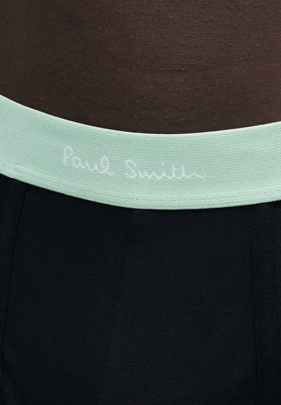Gros plan de la taille d'une personne portant un pantalon noir avec une ceinture vert clair affichant un texte blanc pâle "Paul Smith".