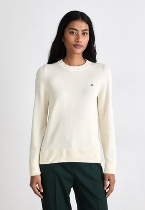 Cream-colored knit sweater with a ribbed neckline and cuffs, featuring a small logo on the chest. Smooth texture and straight fit design.