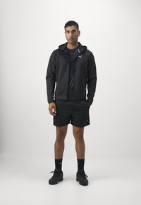 Schwarze Sportjacke mit Kapuze, kombiniert mit schwarzen Shorts und Socken. Merkmale sind das Puma-Logo auf der Jacke, der strukturierte Stoff und Sportschuhe.