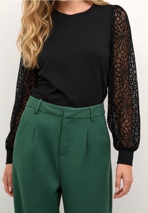 Top nero con maniche lunghe in pizzo trasparente abbinato a pantaloni verdi a vita alta su una persona con capelli biondi lunghi e mossi.