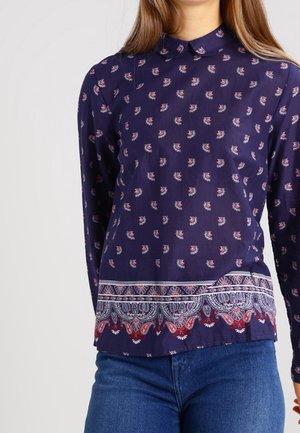 Marineblå top med lange ærmer med små lyserøde blomstertryk og detaljeret hvid og rød paisley kantdesign, kombineret med blå jeans.