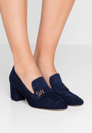 Kvinde iført navy blå ruskinds loafers med blokhæle og guld "SW" logo på stropperne, stående på et glat hvidt gulv.