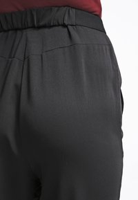 Pantalon en tissu noir lisse avec une taille élastique froncée et des poches latérales, montrant le détail arrière et la légère brillance du matériau.