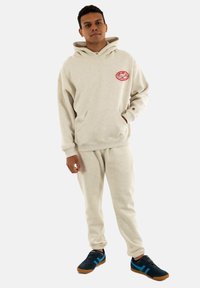 Sudadera de algodón beige con un bolsillo frontal, que presenta un logo rojo. Combinada con pantalones de chándal a juego y zapatillas azules con suelas marrones.