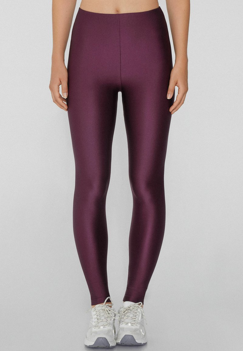 Leggings bordeaux a vita alta realizzati in un tessuto elastico lucido, design aderente, texture liscia, abbinati a scarpe da ginnastica bianche.