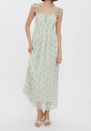 Kobieta w białej, kwiecistej, falbaniastej sukience midi bez rękawów z kwadratowym dekoltem, nosząca beżowe sandały na słupku z paskami.