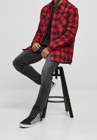 Camisa de franela a cuadros roja y negra, camiseta negra, jeans grises y zapatillas con estampado de cuadros en blanco y negro, sentado en un taburete negro.