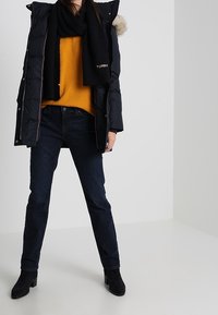 Zwarte gewatteerde jas met een met bont afgewerkte capuchon, gedragen over een oranje sweater en een zwarte sjaal. Gecombineerd met donkere blauwe jeans en zwarte enkellaarsjes.