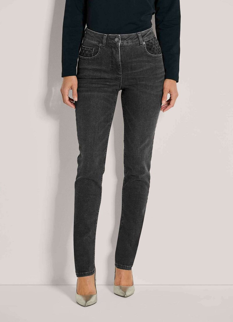 Vrouw in high-waisted zwarte skinny jeans met versierde voorzakken en zilveren puntige hakken tegen een neutrale achtergrond.
