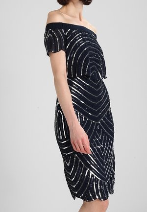 Frau in einem dunklen schulterfreien Kleid mit silbernen Pailletten in geometrischen Mustern und kurzen Ärmeln vor einem einfarbigen hellen Hintergrund.