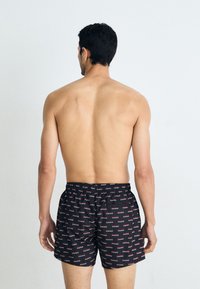 Shorts de baie negre cu un model repetitiv al logo-ului "HUGO" în alb și roșu. Talie elastică și croială clasică. Modelul este văzut din spate.