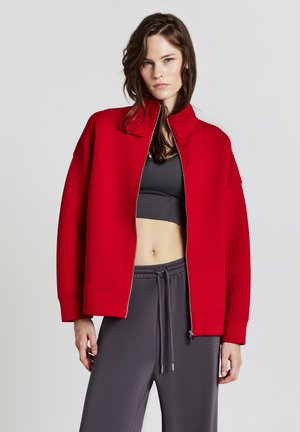 Rote Zip-Jacke mit hohem Kragen, aus weichem Material gefertigt. Verfügt über übergroße Ärmel und einen lässigen Schnitt. Kombiniert mit grauen Hosen.