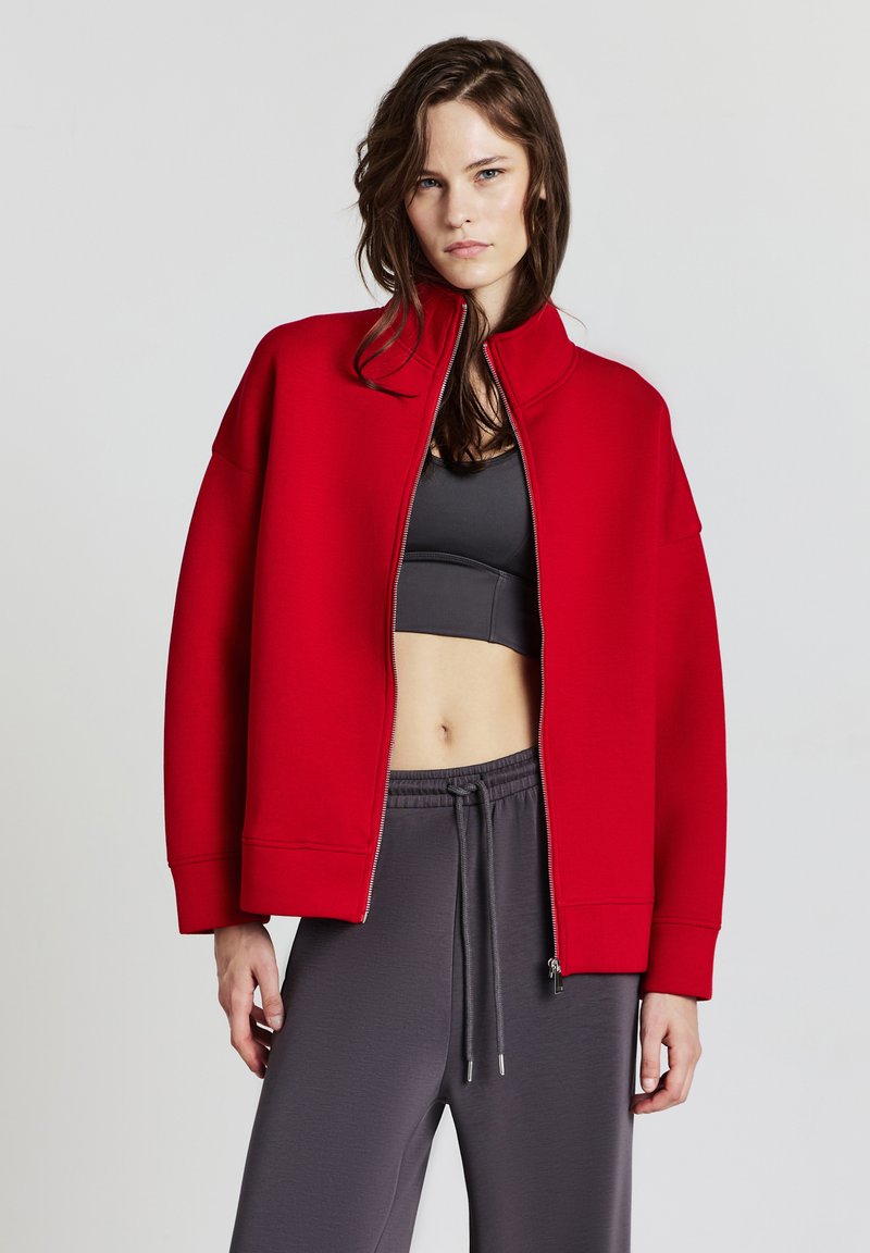 Giacca rossa con zip e colletto alto, realizzata in materiale morbido. Presenta maniche oversize e una vestibilità rilassata. Abbinata a pantaloni grigi.