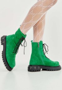 Botas de ante verdes hasta el tobillo con cordones negros, cremallera lateral y suela gruesa negra con acentos de hardware plateado. Diseño texturizado y rugged.