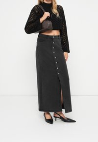 Jupe maxi en denim noir avec une fente à l'avant, ornée de boutons argentés au centre. Associée à un crop top en tricot noir et des talons noirs.