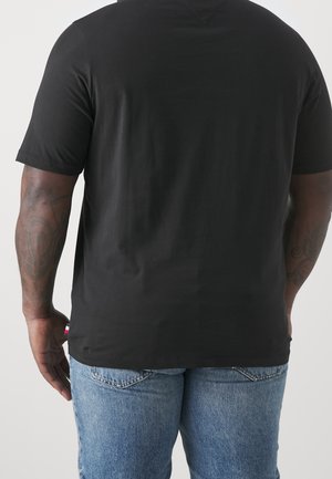 Person iført en ensfarvet sort kortærmet t-shirt og lyseblå denimjeans, vist bagfra med synlige tatoveringer på armene.