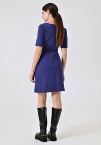 Blauwe jurk met korte mouwen, een aansluitende taille, plooien en een gladde textuur. Gecombineerd met nette zwarte laarzen tot de knie met gouden accenten.