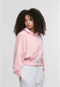 Felpa cropped rosa con cappuccio, maniche ampie e orlo a coste. Presenta un'etichetta sulla vita. Abbinata a pantaloni della tuta grigi chiari.