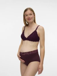 Έγκυος γυναίκα φορώντας μαύρα δαντελένια εσώρουχα εγκυμοσύνης, κρατώντας τρυφερά τη�ν κοιλιά της και χαμογελώντας μπροστά σε λευκό φόντο.