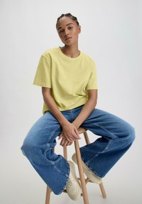 Geel T-shirt met korte mouwen van katoen, gecombineerd met hoge blauwe wijde broek en off-white sneakers, zittend op een houten kruk.