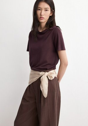 Mlada ženska z dolgimi temnimi lasmi, oblečena v bordo kratko majico s kratkimi rokavi, temno rjave široke hlače in bež pas iz šal povezanim okoli pasu.