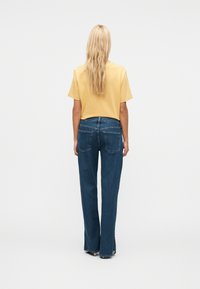 Ξανθιά γυναίκα που στέκεται με την πλάτη, φορώντας κίτρινο πουκάμισο με κοντά μανίκια και σκούρο μπλε τζιν με ίσια μπατζάκια και πίσω τσέπες.