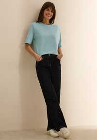 Camicia azzurro chiaro a maniche corte abbinata a jeans scuri a vita alta. Sneakers bianche e una superficie liscia in tutto. Sfondo naturale.