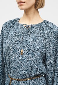 Kobieta ubrana w niebieską, kwiecistą bluzkę z długim rękawem, z wiązaniem przy szyi i złotym paskiem łańcuszkowym w talii, na jednolitym tle.