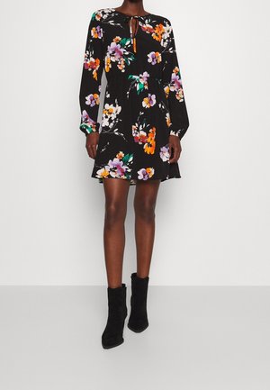 Mujer con un vestido largo de manga larga negro con estampado floral, escote con lazo y botines negros, posando frente a un fondo liso.