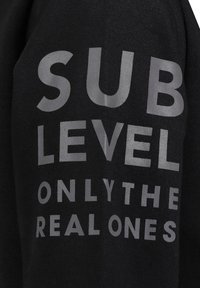 Zwarte trui met vetgrijze tekst: "SUB LEVEL ONLY THE REAL ONES." Gemaakt van zachte stof met een relaxte pasvorm.