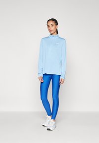 Maglietta atletica a maniche lunghe di colore azzurro chiaro con zip a quarto, dal tessuto morbido, abbinata a leggings blu lucidi e sneakers bianche.