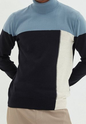 Mand iført en farveblokeret rullekravetrøje med blå, sort og hvide sektioner kombineret med beige fløjlsbukser.