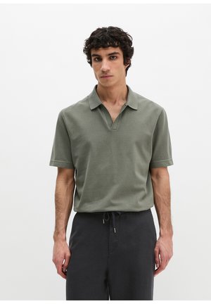 Mann mit lockigem Haar, der ein olivgrünes Polohemd mit kurzen Ärmeln und dunkle Hose mit Kordelzug trägt, vor einem schlichten weißen Hintergrund stehendes.