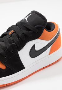 Sneaker nera, arancione e bianca con texture in pelle e camoscio. Presenta una suola in gomma, lacci e logo ricamato sulla linguetta.