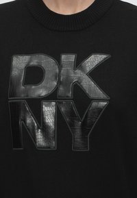 Pull noir avec de grandes lettres noires brillantes en relief "DKNY" sur la poitrine, avec un motif texturé ressemblant à une peau de reptile.