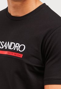 Camiseta negra de cuello redondo con texto bordado en blanco y rojo "SANDRO" y "ZAVETTI MILANO" en el pecho, llevada por una persona.