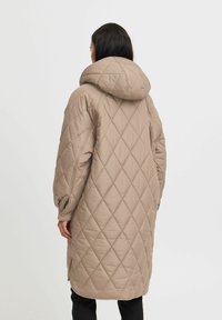 Manteau long en beige matelassé avec une capuche, présentant des motifs en diamant, des poignets élastiques et des fentes latérales pour un plus grand confort de mouvement.